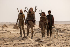 EXCLUSIVE: “The Wheel Of Time” Costume Designer Sharon Gilham Talks Season 3 3 (Left to right) Maja Simonsen as Chiad, Ragga Ragnars as Bain, Ayoola Smart as Aviendha, and Marcus Rutherford as Perrin Aybara, walking through a desert. Aviendha, Bain and Chiad all have long reddish-brown hair. They wears very similar outfits: sand-brown hoods, black scarves, brown leather armor over sand-brown tunics, skirts and trousers, with short spears strapped to their backs. Perrin has short dark frizzy hair and a beard, and wears a dark green woven-leather vest over a faded red long-sleeved shirt with olive-green trousers and brown boots.