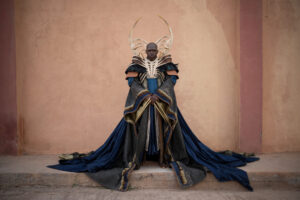 EXCLUSIVE: “The Wheel Of Time” Costume Designer Sharon Gilham Talks Season 3 4 Daniel Francis as High Lord Turak, standing in front of a pink stucco wall. He is bald. He wears an intricate collar of bone over a black feathered mantle, over a black-and-gold scale-patterned robe with a long dark blue train. The first two fingernails on both his hands are extremely long and bladed.