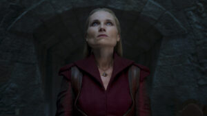 EXCLUSIVE: “The Wheel Of Time” Costume Designer Sharon Gilham Talks Season 3 6 Kate Fleetwood as Liandrin Guirale, standing in a stone doorway, looking up. She has long blonde hair. She wears a red-brown leather vest over a long-sleeved red coat, with a red stone hanging around her neck.